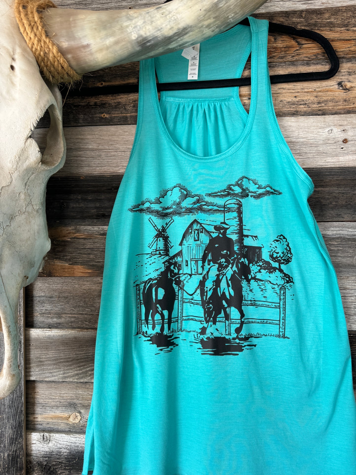 Blue tank top hanging on a hanger decorated with a black screenprint transfer from Snappy 6 Co that has a cowboy and a barn and windmill on it 