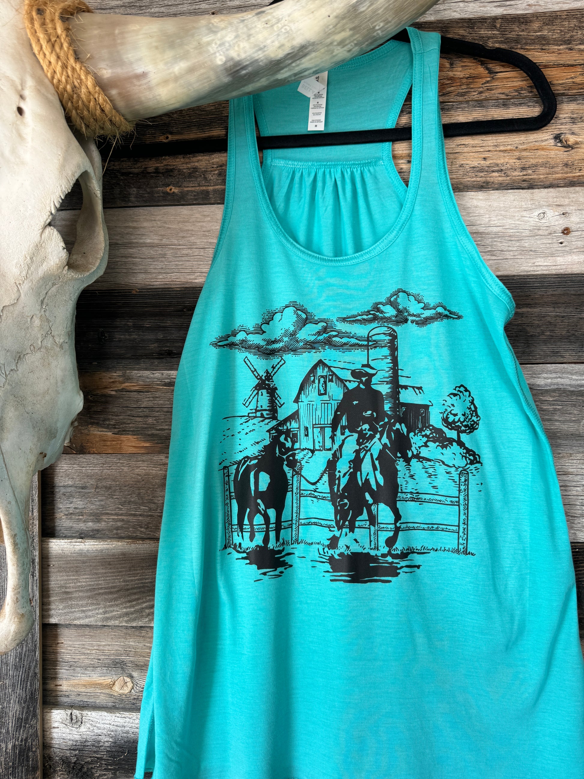 Blue tank top hanging on a hanger decorated with a black screenprint transfer from Snappy 6 Co that has a cowboy and a barn and windmill on it 